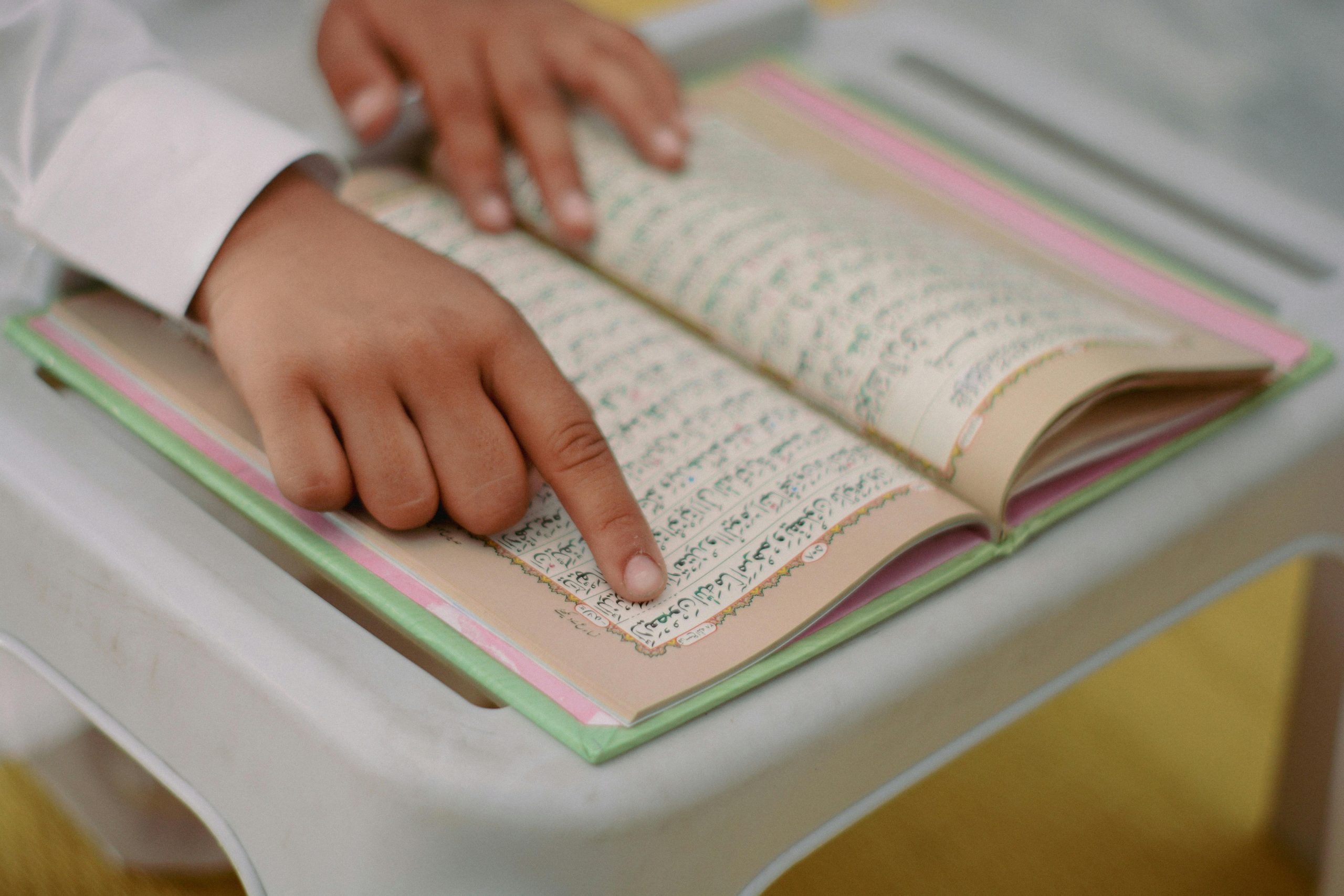 A close-up of a child reading the Quran, highlighting a finger tracing words on a desk indoors.